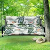 A swing seat cushion with a tropical leaf pattern is placed on a swing in a garden setting. Produktbild-Vorschau