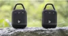 Two black portable Bluetooth speakers are placed on a rock with a blurred green background. Voir les images du produit