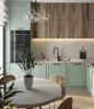 A modern kitchen with light green cabinets, wooden upper cabinets, and a white round dining table in the foreground. Produktbild-Vorschau