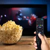 A hand holding a remote control in front of a television screen with a bowl of popcorn. Produktbild-Vorschau