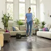 A woman is steam cleaning a wooden floor in a bright, modern living room. The room has large windows, sofas, and potted plants. Podgląd zdjęcia produktu