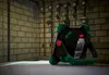 A portable work light is placed on a floor in front of a brick wall with pipes and gauges. Produktbild-Vorschau