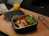 A heated lunchbox filled with food, placed on a wooden surface next to a glass of orange juice and cutlery. Produktbild-Vorschau
