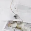 A close-up view of a beige roller blind with a decorative embroidered wave pattern and a metal eyelet. The blind is attached to a window frame using clips, and there are images of jars and a bowl of white flakes visible behind it. Produktbild-Vorschau