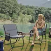 Two camping chairs are set up outdoors by a lake with trees in the background. A woman is sitting in one of the chairs. Anteprima dell’immagine prodotto