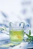A clear glass mug filled with green tea, placed on a white cloth with some sprigs of linden flowers. Produktbild-Vorschau