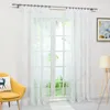 A white sheer curtain with a leaf pattern hangs in front of a window, with a yellow chair and a lamp visible. Produktbild-Vorschau