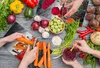 A top-down view of a collection of fresh vegetables and hands preparing food on a wooden table. Produktbild-Vorschau