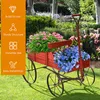 A red wooden wagon filled with pink and blue flowers sits on a green lawn in a garden setting. Produktbild-Vorschau