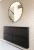 A black shoe cabinet with four drawers is mounted on a wall with patterned wallpaper. A round gold mirror hangs above the cabinet. Produktbild-Vorschau