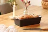 A loaf of bread in a baking dish on a wooden table, with a candle and flowers in the background. Produktbild-Vorschau