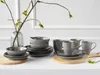 A set of grey stoneware dishes and mugs are arranged on a white surface with a vase of branches in the background. Náhľad obrázku produktu