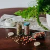 Small glass jars filled with seeds and nuts, along with a jar of yogurt, are arranged on a wooden table next to a potted fern. Anteprima dell’immagine prodotto