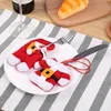 Christmas-themed cutlery holders are placed on a white plate on a striped placemat, with a wine glass and bread in the background. Produktbild-Vorschau
