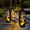 Two solar-powered garden lights with a tree design are placed on a stone pathway in a garden at dusk. Produktbild-Vorschau
