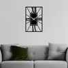 A modern black metal wall clock with Roman numerals is mounted on a light gray wall above a gray sofa with two pillows. Produktbild-Vorschau