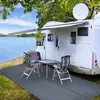 A campsite scene with a motorhome, awning, outdoor furniture, and a mat by a lake. Produktbild-Vorschau