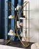 A gold metal bookshelf with glass shelves, decorated with various vases and books, set against a blue curtain. Náhľad obrázku produktu
