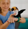 A child's hand wearing a toucan toy on their wrist, with the child's face blurred in the background. Voir les images du produit