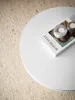 A white round coffee table with a book and two candle holders on it, placed on a textured rug. Náhľad obrázku produktu