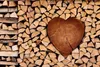 A heart-shaped piece of wood is placed in the center of a neatly stacked pile of firewood. Produktbild-Vorschau