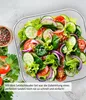 A close-up overhead view of a fresh salad in a clear bowl, with ingredients like lettuce, tomatoes, cucumbers, and red onions visible. A small wooden bowl of peppercorns is in the upper left corner. Produktbild-Vorschau