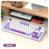 A white gaming keyboard with purple and light purple keycaps is shown on a desk with books and a Nintendo Switch. Produktbild-Vorschau