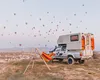 A camper van is parked on a hill with a hammock set up in front of it. Many hot air balloons are visible in the sky. Podgląd zdjęcia produktu