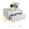 A white floating nightstand with an open drawer, displaying a book, a decorative lamp, and a vase with flowers on top. A power cord with a switch is visible. Produktbild-Vorschau