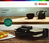 A Bosch contact grill is shown on a wooden surface, with a sandwich on a plate in the foreground and two kitchen accessories in the background. Podgląd zdjęcia produktu