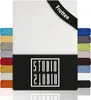 A white fitted sheet with a black band at the bottom displaying the brand logo. Several swatches of different colored fitted sheets are stacked on the left and right sides. Produktbild-Vorschau