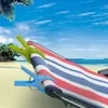 Two large plastic clips holding a striped beach towel on a beach with palm trees and the ocean in the background. Produktbild-Vorschau