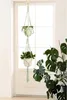 A macrame plant hanger with two potted plants hanging in front of a window, with a large monstera plant in the foreground. Produktbild-Vorschau