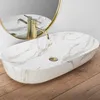 A marble-patterned oval sink with a gold faucet, set against a white wall and a gold-rimmed mirror, on a wooden floor. Náhľad obrázku produktu