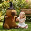 A young girl is sitting on the grass in a garden next to a large stuffed capybara wearing a wizard hat and cape. Náhľad obrázku produktu