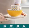 A person is holding a stack of yellow and white patterned ceramic dishes, including plates, a bowl, and a mug. Náhled obrázku produktu