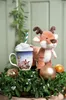 A plush reindeer toy sits next to a mug of hot chocolate, surrounded by festive greenery and ornaments. Produktbild-Vorschau