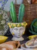 A close-up of a decorative ceramic pot shaped like a face, adorned with lemons and leaves, holding two green cacti. The pot is placed on a dark surface, with other decorative items like plates and fabric visible in the background. Náhľad obrázku produktu