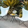 Two white butterfly chairs and a small round table with a glass top are set up on a patio area with lush green and yellow foliage in the background. Náhľad obrázku produktu