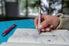 A person's hand is writing in a notebook with a pink pen, with another pink pen on a blue cutting mat. Produktbild-Vorschau