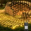 A net of warm white fairy lights is draped over large rocks and spread across a grassy lawn, with a power adapter visible in the foreground. Náhled obrázku produktu