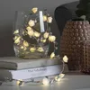 A string of LED lights shaped like roses is placed inside a glass vase, which is sitting on a stack of books. Another decorative vase is visible in the background. Produktbild-Vorschau