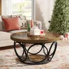 A round coffee table with a wood top and black metal base, decorated for Christmas with small trees, a house figurine, and a candle. A sofa and a Christmas tree are visible in the background. Anteprima dell’immagine prodotto
