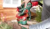 A person is holding a Bosch paint sprayer outdoors, with a wooden building and plants in the background. Produktbild-Vorschau