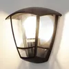 A black outdoor wall lamp with a clear glass shade is mounted on a white wall. Produktbild-Vorschau