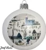 A white Christmas ornament with a painted winter village scene. The scene depicts a church on a hill, houses, trees, and two people pulling a sled in the snow. Náhled obrázku produktu