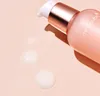 A close-up shot of a pink cosmetic pump bottle with rose gold accents, with three drops of serum on a light pink surface. Podgląd zdjęcia produktu