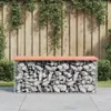 A gabion bench with a wooden top sits on a paved patio in a garden setting, with a wooden fence and greenery in the background. Náhled obrázku produktu