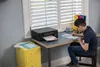 A young boy is sitting at a desk, using a printer and writing on a piece of paper. The scene is set in a room with a window and blinds. Podgląd zdjęcia produktu
