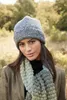 A woman wearing a knitted hat and scarf, outdoors in a natural setting. Produktbild-Vorschau